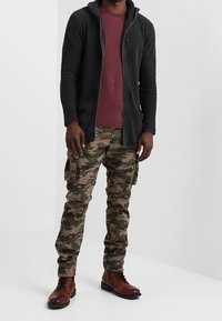 Cardigan en maille noire à capuche, t-shirt bordeaux, pantalon cargo camouflage et bottines marron à lacets. Tissu texturé et coupe décontractée détaillés.