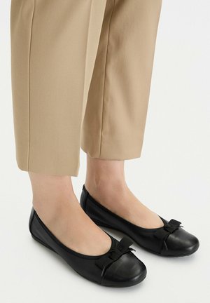 Pies con pantalones cortos beige y bailarinas de cuero negras con lazos de tela sobre un fondo blanco.