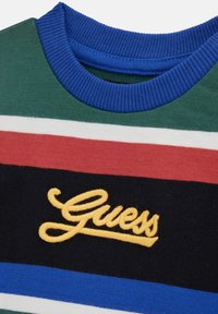 Maglione a righe multicolori con sezioni verdi, rosse, bianche, blu e nere. Logo "Guess" ricamato giallo sulla parte anteriore. Collo a coste blu.