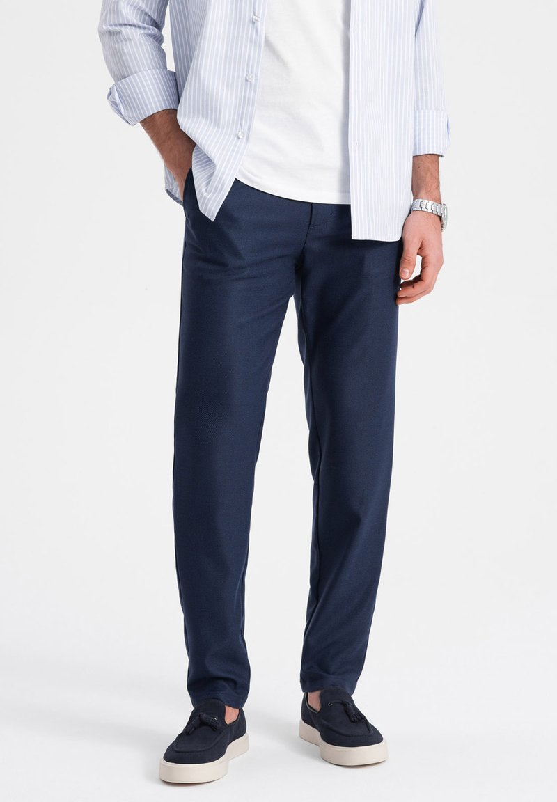Pantalons bleu marine en tissu lisse et texturé, offrant une coupe décontractée et des poches latérales, associés à des baskets bleu marine et à une chemise à rayures claires.