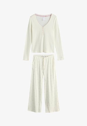 Bluză lungă cu mâneci lungi, cu nervuri, de culoare crem, cu decolteu în V și doi nasturi, asortată cu pantaloni largi crem cu un motiv mic floral portocaliu.