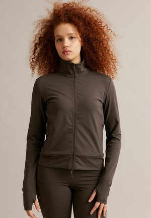 Junge Frau mit lockigem roten Haar, die eine dunkelbraune Jacke mit Reißverschluss und passende Hose vor neutralem Hintergrund trägt.