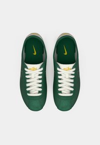 Une paire de baskets Nike vertes avec des lacets blancs, des logos Nike jaunes sur la languette et les semelles intérieures, sur un fond blanc uni.