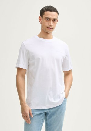 Weißes Baumwoll-T-Shirt mit runder Ausschnitt, kurzen Ärmeln und dezentem Logo-Detail auf der Brust. Getragen mit hellblauen Jeans.