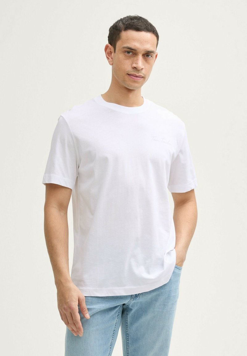 Hvid bomulds t-shirt med rund halsudskæring, korte ærmer og diskret logo detalje på brystet. Båret med lyseblå denim jeans.