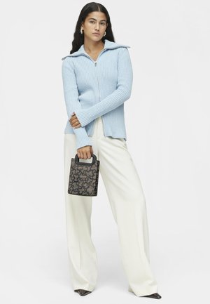 Pullover en maille côtelée bleu clair avec un col montant, fermeture éclair et manches longues. Le mannequin tient un sac à main noir à motif avec une poignée.