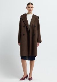 Brauner Wollüberrock mit großem, spitzem Kragen, doppelt gereihten schwarzen Knöpfen und weit geschnittenen Ärmeln; kombiniert mit einem weißen Oberteil und einem dunklen Rock.