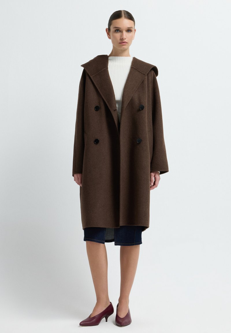 Brauner Wollüberrock mit großem, spitzem Kragen, doppelt gereihten schwarzen Knöpfen und weit geschnittenen Ärmeln; kombiniert mit einem weißen Oberteil und einem dunklen Rock.