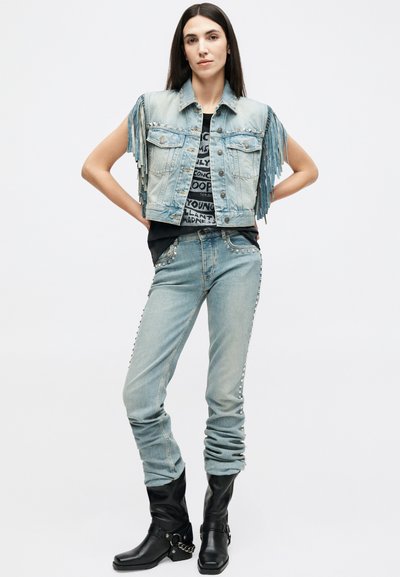 Γυναίκα που φοράει γαλάζιο τζιν γιλέκο με κρόσσια, μαύρο μπλουζάκι με γραφικά, αντίστοιχο τζιν παντελόνι με τρουκς και μαύρα καουμπόικα μποτάκια, με τα χέρια στη μέση.