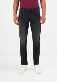 Mann in engen, ausgewaschenen schwarzen Jeans, weißen Sneakers und einem burgunderroten Hemd, der vor einem schlichten weißen Hintergrund steht.