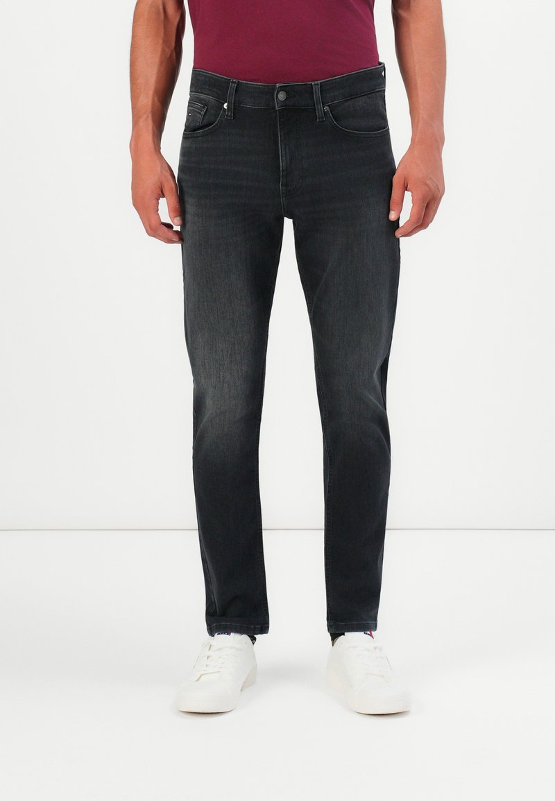 Mann in engen, ausgewaschenen schwarzen Jeans, weißen Sneakers und einem burgunderroten Hemd, der vor einem schlichten weißen Hintergrund steht.