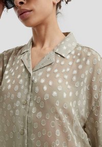 Donna che indossa una blusa traslucida beige con un motivo circolare testurizzato, con bottoni sul davanti e colletto aperto, mostrata dalla gola al torace.