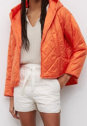 Kvinde iført en orange quiltet jakke over en hvid striktrøje og hvide quiltede shorts med snøre i taljen.