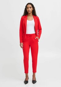 Rode op maat gemaakte pak met een blazer en verstelbare trekkoordbroek. Gladde stof, slim fit en zwarte hoge hakken maken de look compleet.