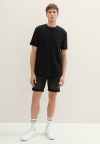 Czarna koszulka z krótkim rękawem z kieszonką na piersi, w połączeniu z podwiniętymi dżinsowymi szortami; białe sneakersy i paskowane skarpety dopełniają strój.