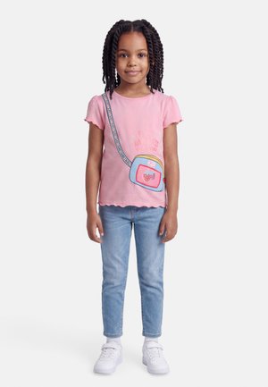 Νεαρή κοπέλα με πλεγμένα μαλλιά, ντυμένη με ροζ μπλούζα με στάμπα τσάντας, μπλε τζιν και άσπρα αθλητικά παπούτσια, στέκεται μπροστά σε ένα απλό φόντο.