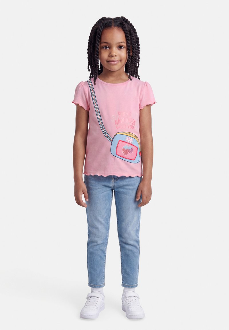 Νεαρή κοπέλα με πλεγμένα μαλλιά, ντυμένη με ροζ μπλούζα με στάμπα τσάντας, μπλε τζιν και άσπρα αθλητικά παπούτσια, στέκεται μπροστά σε ένα απλό φόντο.