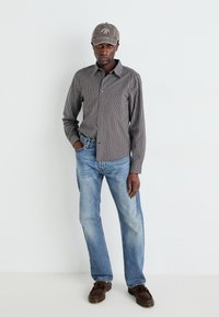 Checkered button-up shirt in black and white, blue jeans, brown loafers, and gray baseball cap with logo. Features long sleeves and relaxed fit.