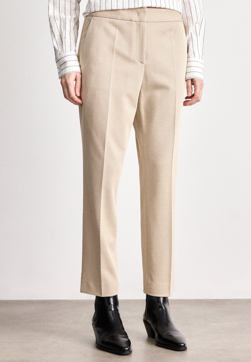 Calças beige ajustadas com um tecido texturizado, corte reto e costuras laterais. Combinadas com botas de tornozelo pretas.