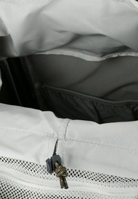 Interior de una bolsa de tela gris claro con un bolsillo de malla; presenta un cierre con un tirador oscuro. Textura suave y ligeramente brillante.