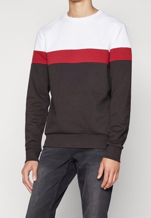 Homme portant un sweat à blocs de couleurs avec un panneau supérieur blanc, une bande rouge et le bas marron foncé, associé à un jean gris foncé.