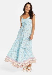 Light blue maxi dress with floral patterns, spaghetti straps, tiered ruffles, and decorative trim at the hem; worn with platform sandals.