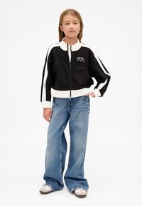 Jovem em pé vestindo uma jaqueta preta e branca com fecho éclair, jeans azuis de perna larga e ténis brancos contra um fundo simples.