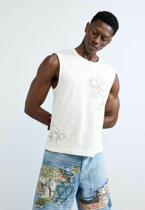 Hombre musculoso vistiendo una camiseta blanca sin mangas con bordado floral y shorts de mezclilla con parches, posando contra un fondo claro y liso.