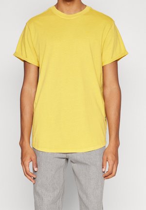 T-shirt en coton jaune à manches courtes, avec un col rond et des revers aux manches, porté par une personne portant un jean gris.