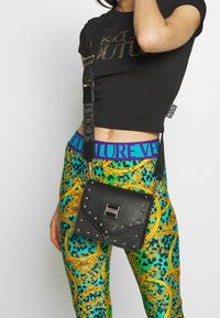 Borsa a tracolla in pelle nera con borchie argentate, patta triangolare e logo dorato. La tracolla attaccata presenta l'etichetta "COUTURE" in lettere in grassetto.