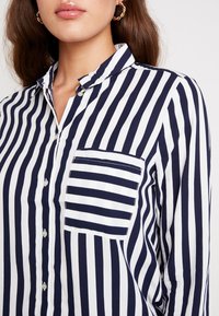 Chemise à rayures bleu marine et blanches avec boutons, dotée d'un col, d'une poche avant et de manches longues. Le tissu est lisse avec une légère brillance.