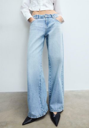 Personne portant un jean large bleu clair avec les mains dans les poches et des chaussures noires pointues, debout contre un mur blanc.