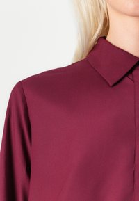 Burgundy blouse with a collar, made of smooth fabric, featuring long sleeves and a clean, tailored design. No patterns or accents noted.
