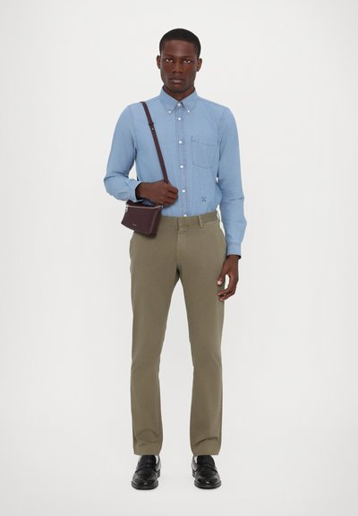 Chemise bleu clair à manches longues avec une poche poitrine, pantalon olive, et un petit sac en bandoulière marron. Des chaussures noires complètent le look.