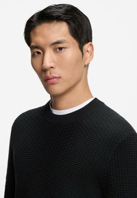 Czarny sweter z dzianiny o teksturowej fakturze z okrągłym dekoltem, założony na białą koszulę z kołnierzykiem. Posiada prążkowany kołnierz oraz dopasowany fason.