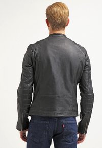 Ein blondhaariger Mann von hinten zu sehen, der eine schwarze Lederjacke mit gepolsterten Ellenbogenpatches und dunkelblaue Jeans trägt.