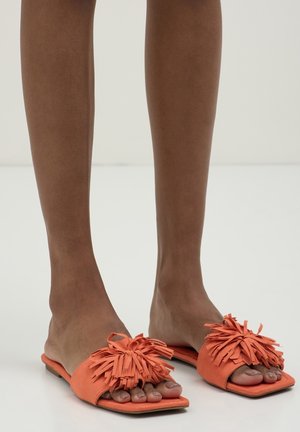 Füße, die orangefarbene Pantoffeln mit dekorativem Fransenbesatz oben tragen und auf einer weißen Oberfläche stehen.