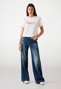 Tricou din bumbac roz cu logo-ul "Guess" în argintiu și roșu, asortat cu blugi largi decolorati de culoare albastră. Modelul stă cu o mână în buzunar.