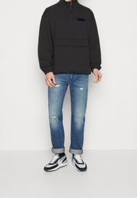 Suéter polar negro con media cremallera, bolsillo delantero y logo bordado; combinado con jeans azul claro desgastados y zapatillas negras con blanco.