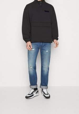 Suéter polar negro con media cremallera, bolsillo delantero y logo bordado; combinado con jeans azul claro desgastados y zapatillas negras con blanco.