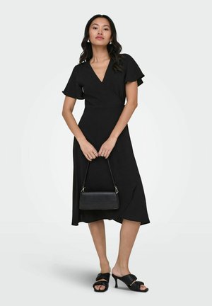 Femme portant une robe midi noire à col en V avec des manches volantées, tenant un petit sac à main noir et portant des sandales noires à talons.
