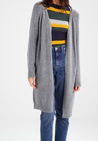 Ljusgrå stickad cardigan med öppen framkant, långa ärmar och ribbade muddar. Bärs över en flerfärgad randig tröja och högmidjade jeans.