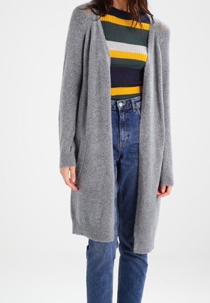 Cardigan gris largo con un ajuste holgado, puños acanalados y un frente abierto, superpuesto sobre un suéter de rayas multicolores y pantalones ajustados de alta cintura.
