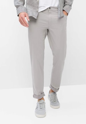 Uomo con pantaloni grigio chiaro risvoltati, sneaker grigie, t-shirt bianca e camicia grigia sbottonata, in piedi con una mano in tasca.