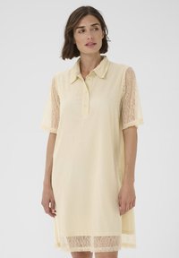 Frau in einem cremefarbenen Spitzenkleid mit kurzen Ärmeln und einem knöpfbaren Kragen, die leicht nach links blickt, vor einem schlichten Hintergrund.