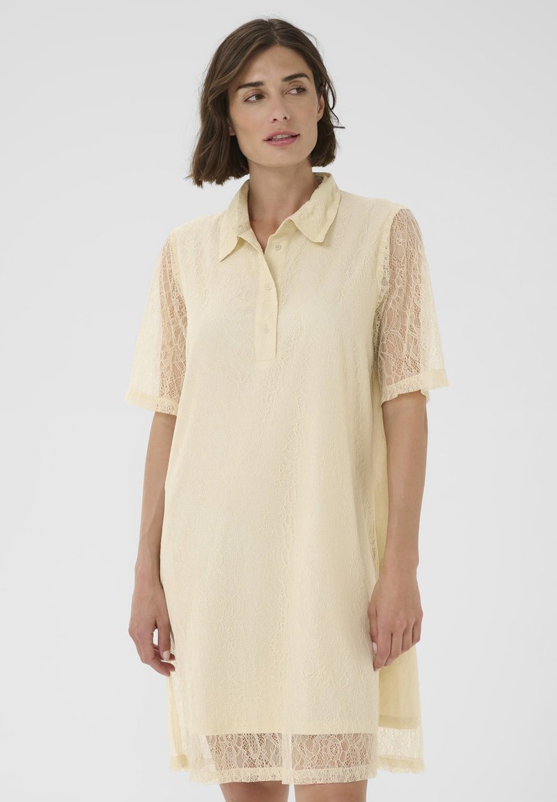 Frau in einem cremefarbenen Spitzenkleid mit kurzen Ärmeln und einem knöpfbaren Kragen, die leicht nach links blickt, vor einem schlichten Hintergrund.