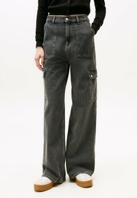 Pantalones cargo en denim gris oscuro, con piernas anchas, bolsillos frontales y detalles de botones a presión. Combinados con una camiseta de manga larga negra y zapatillas blancas.