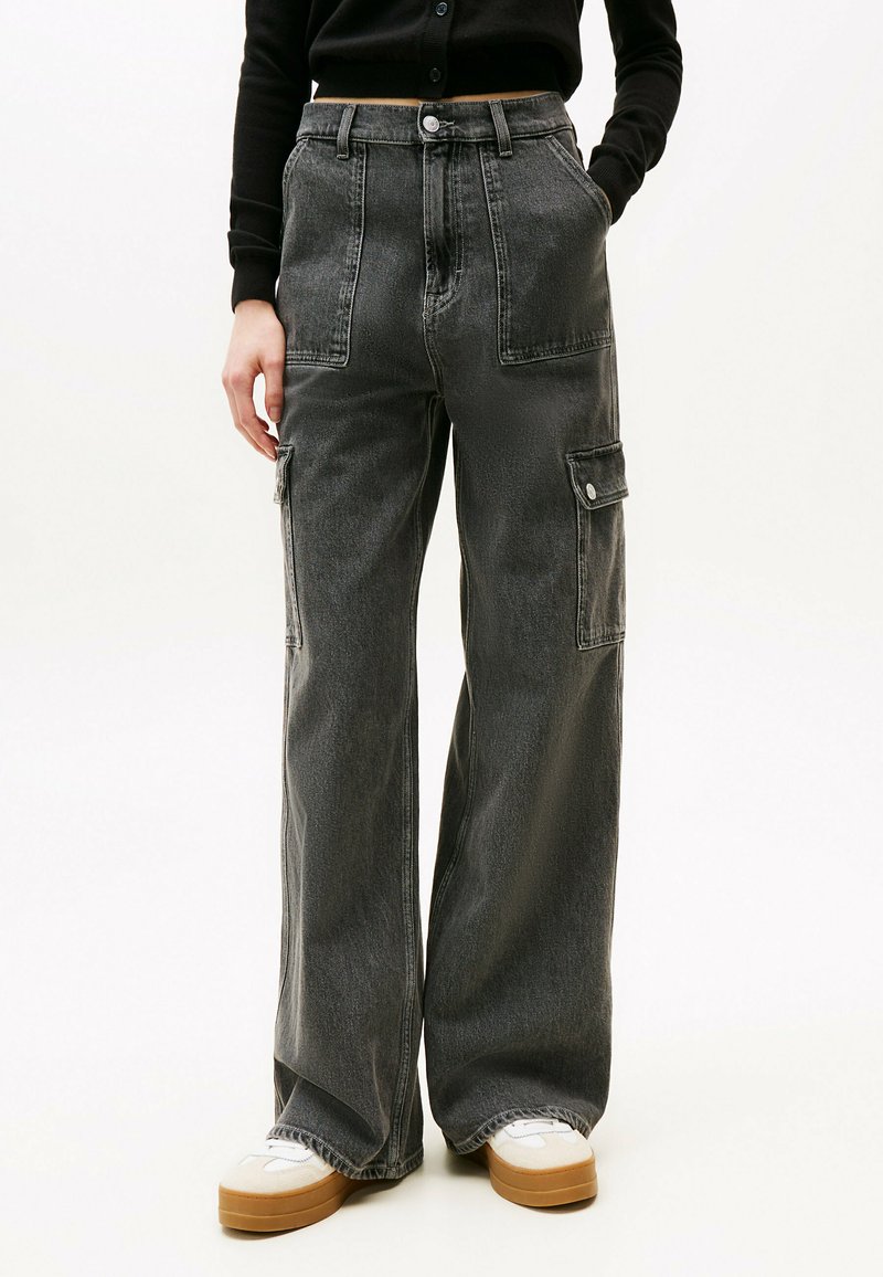Pantalones cargo en denim gris oscuro, con piernas anchas, bolsillos frontales y detalles de botones a presión. Combinados con una camiseta de manga larga negra y zapatillas blancas.