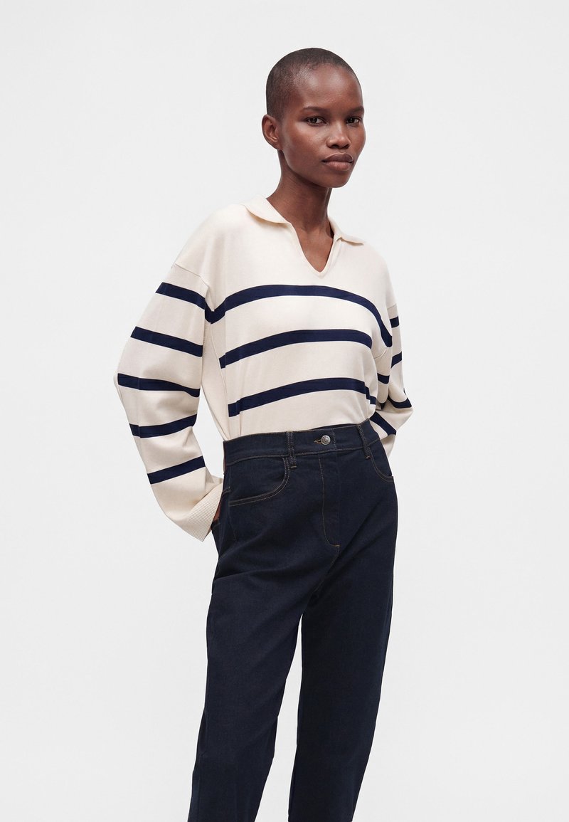 Personne portant un pull crème à rayures bleu marine et un pantalon sombre taille haute, debout devant un fond blanc uni.