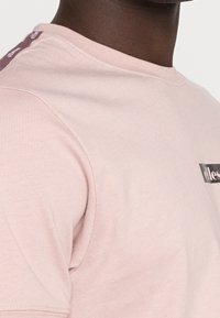 Lyserød T-shirt med rund hals, med et sort logo trykt på forsiden og lilla kantstikning på skulderen.